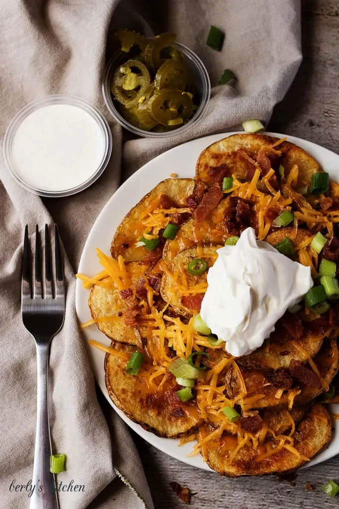 Top down view of sliced potato nachos with cheese, bacon, and a side of ranch and jalapenos.