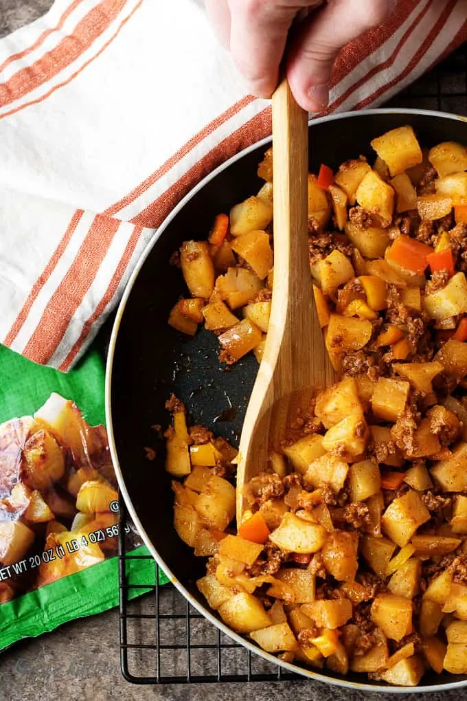 A photo of the potatoes, chorizo, and peppers being cooked in a non-stick skillet. The dish is being stirred with a wooden spoon.