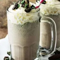 Two oreo milkshakes in chilled mugs with whipped cream and cookies toppings.