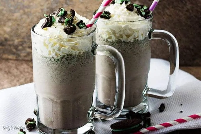 A side-view of two oreo milkshakes in mugs, topped with whipped cream, and crumbled cookies.