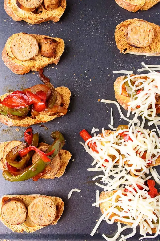 The sausage and peppers crostini bites on a sheet pan being topped with mozzarella to bake.