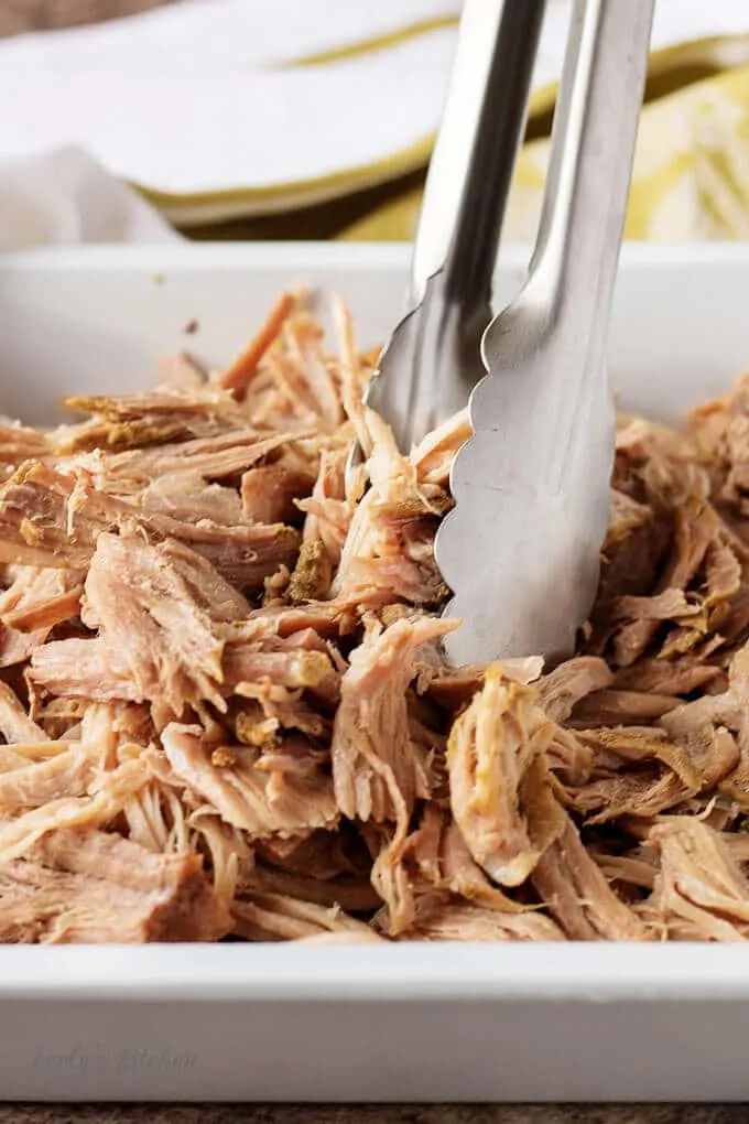 Shredded pieces of pork pot roast in a pan with tongs.