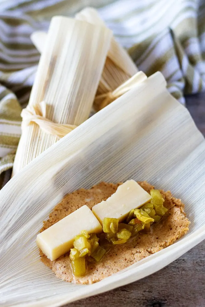 Open corn husk with masa filling, cheese, and green chiles next to closed tamales.