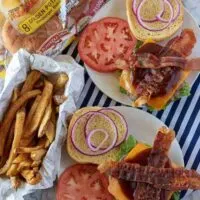 A top-down view of the bacon cheeseburgers being assembled with tomatoes, red onion, and bbq sauce.