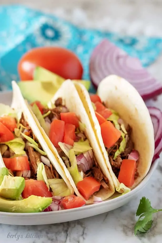 Three instant pot shredded beef tacos on a plate next to a tomato and purple onion.