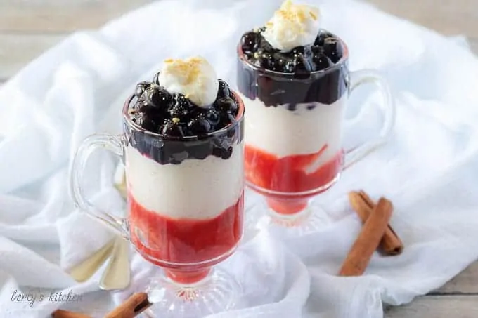 The finished blueberry cheesecake parfaits in mugs layered with strawberries, cheesecake, and blueberry toppings.