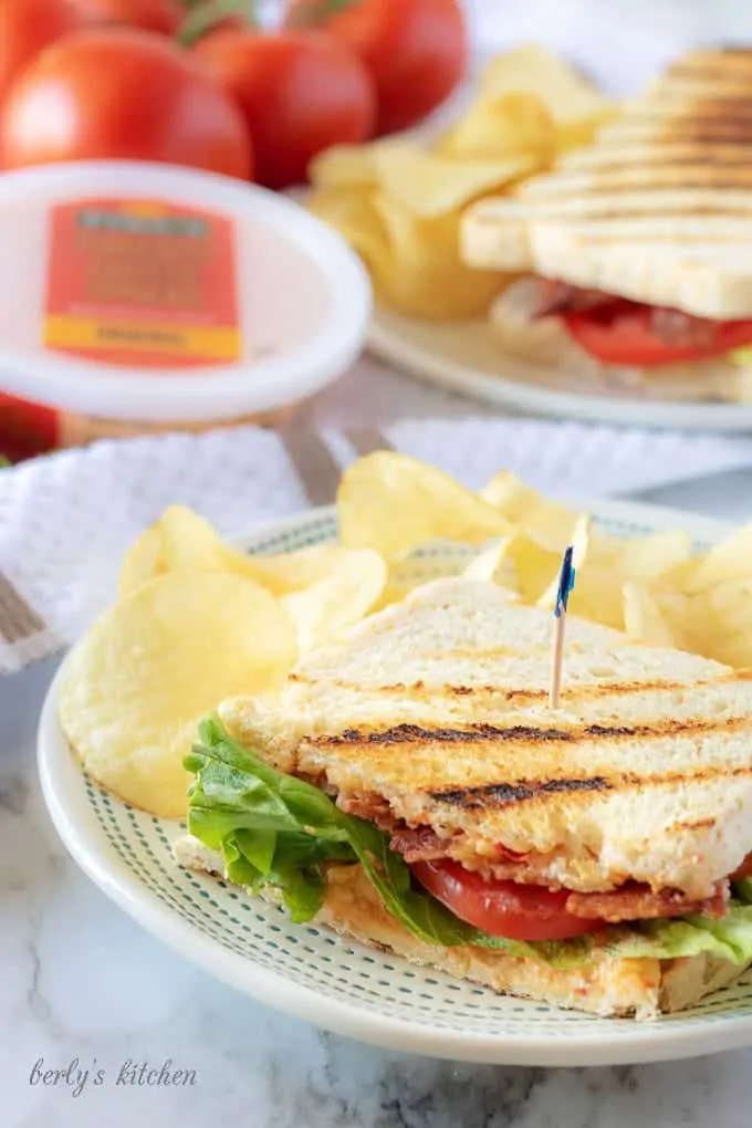 A final photo showing half a pimento cheese sandwich on white plate with a blue tipped toothpick and served with crispy potato chips.