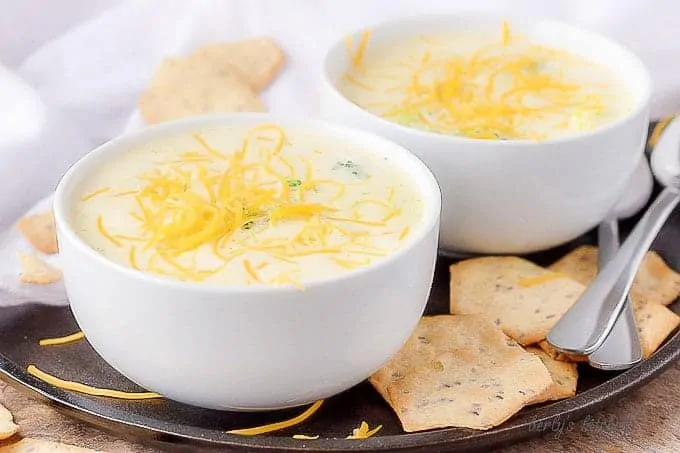 Two bowls of broccoli cheese soup on a flat plate with spoons and crackers.