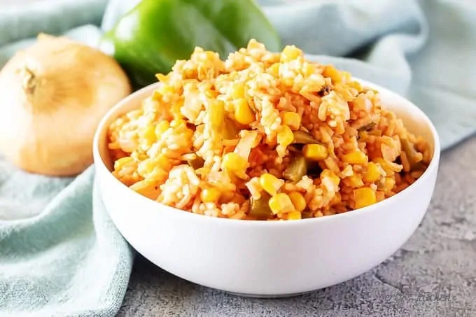 A white bowl of mexican rice surrounded by fresh bell peppers and onions.