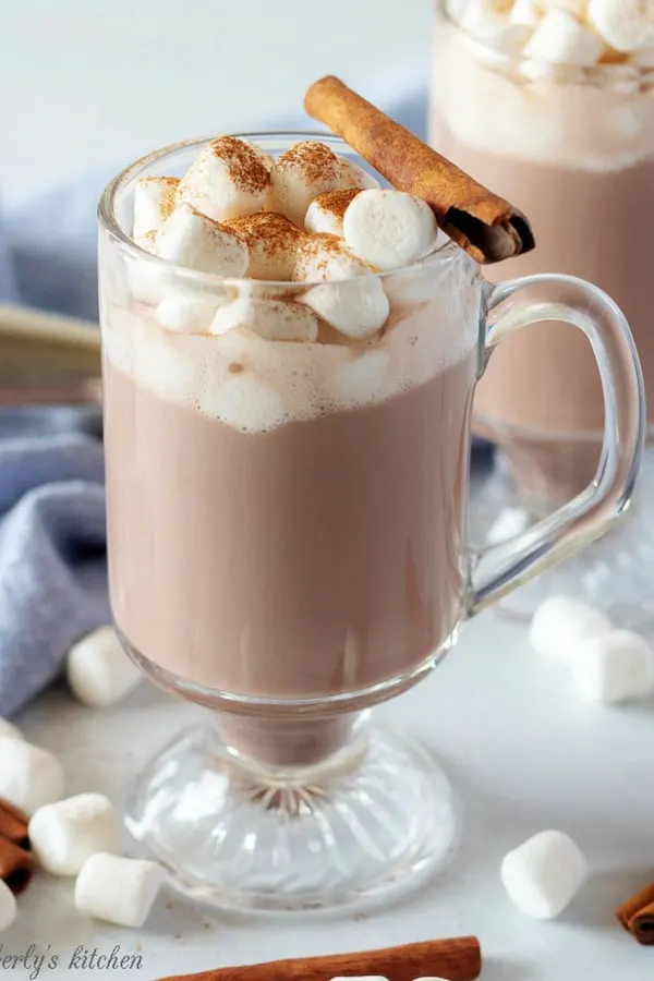 A large photo of the spiked hot chocolate in a clear mug, topped with marshmallows, and garnished with fresh cinnamon and a cinnamon stick.