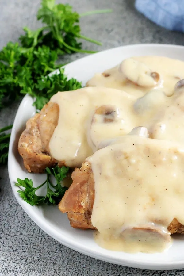 A large photo of the finished instant pot pork chops on a white serving dish, smoothered with mushroom gravy, and garnished with fresh parsley.