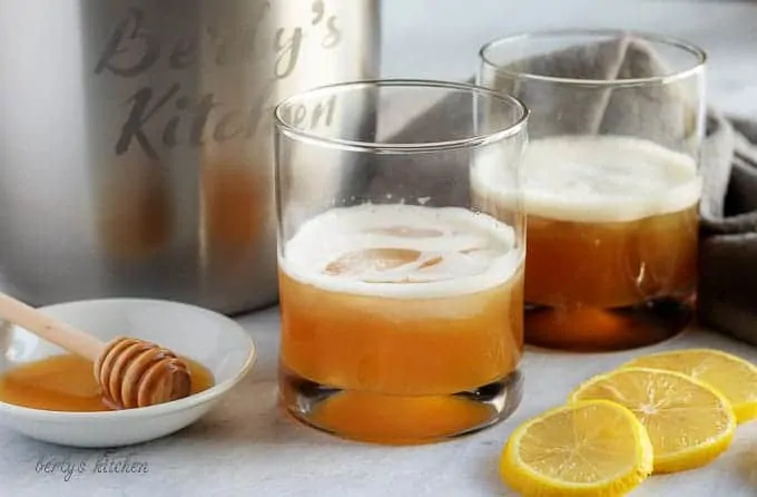 Two gold rush cocktails sitting in a cocktails glasses surrounded by lemon slices and honey on a plate.