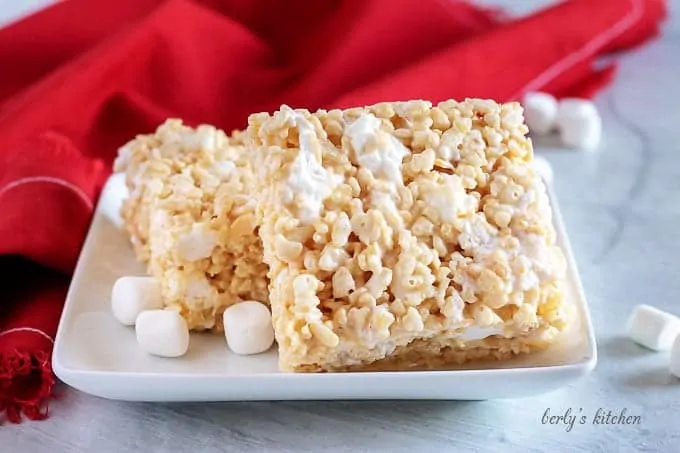 Two perfect rice krispie treats on a square plate next to marshmallows and a red napkin.