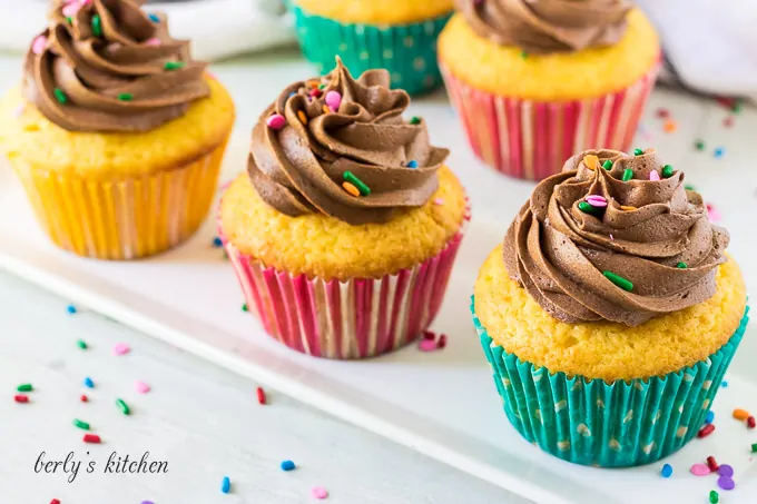 Five cupcakes topped with chocolate buttercream frosting and colorful sprinkles.