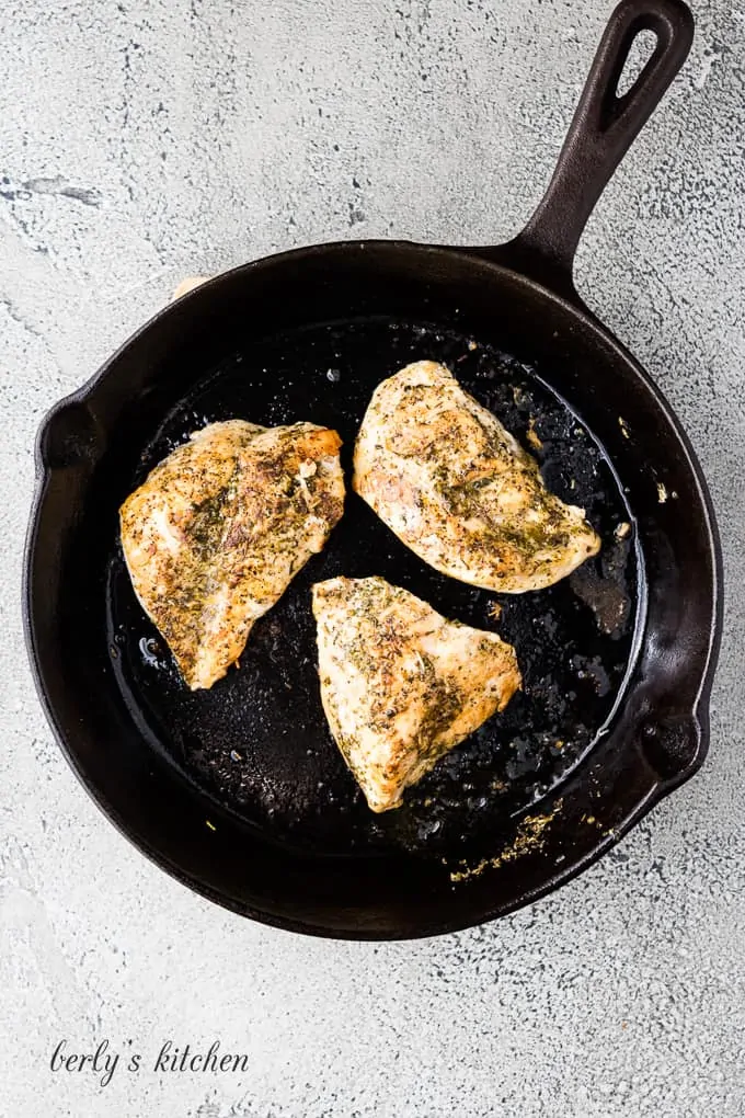 Three seasoned chicken breasts cooking in a cast iron skillet.