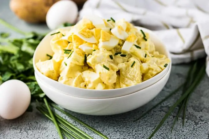 White bowl filled with potato salad next to eggs and chives.