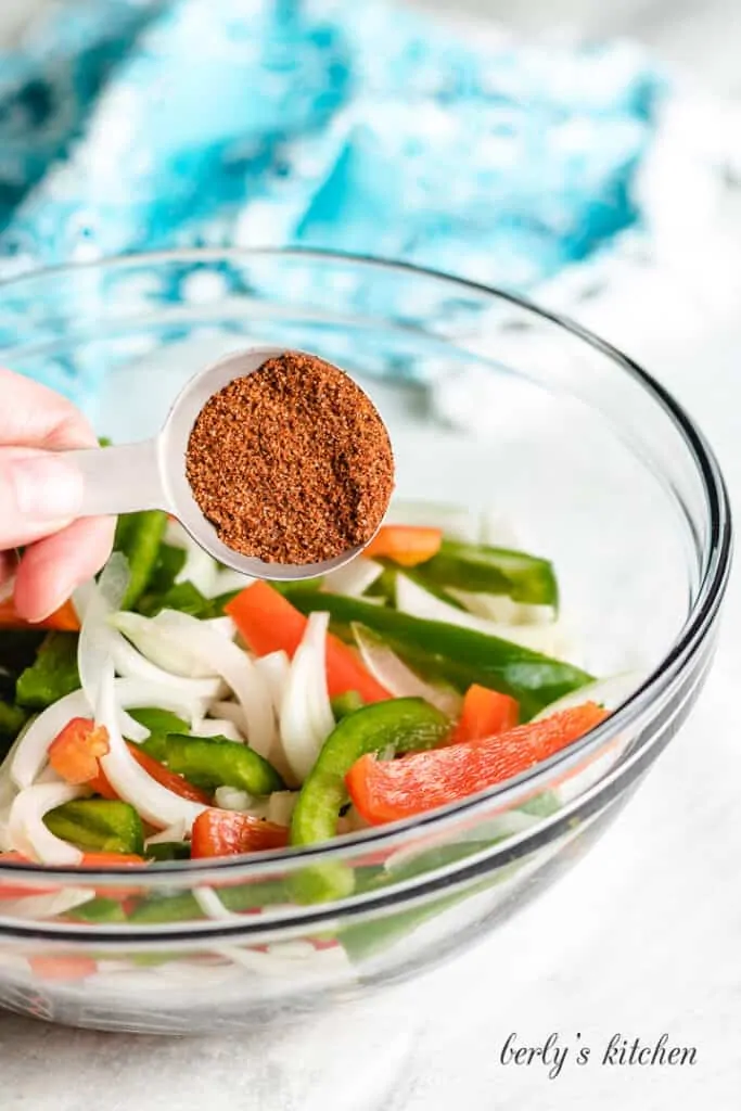 Fajita seasoning being sprinkled over a bowl of sliced peppers and onions.