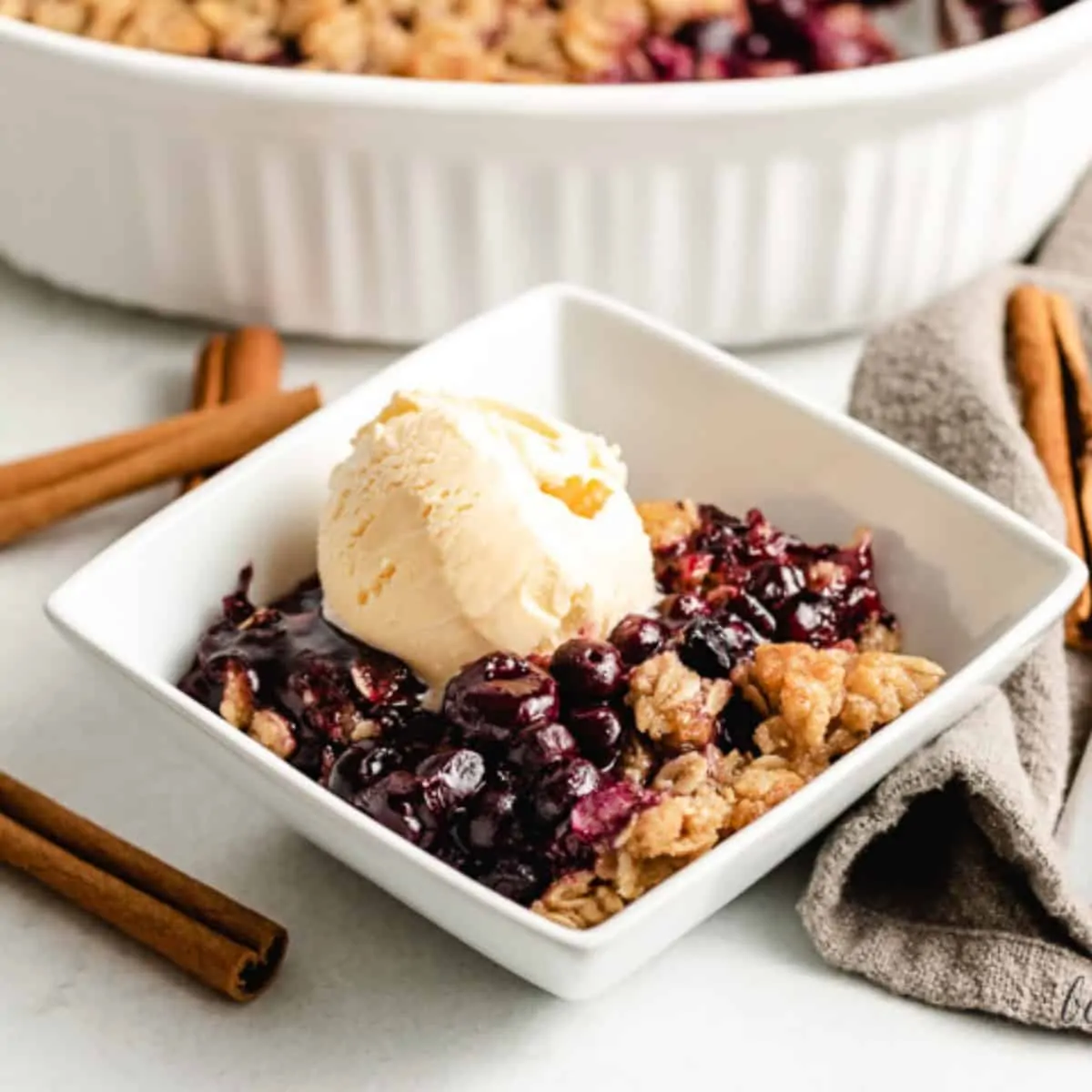 A square bowl with blueberry crisp topped with ice cream.