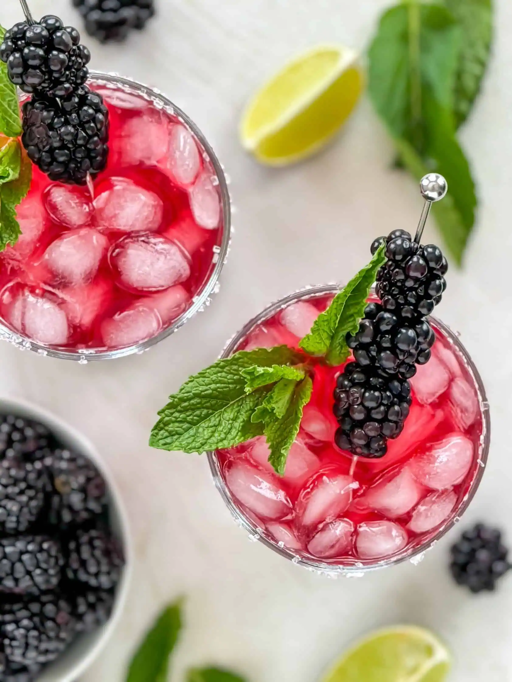 Overhead shot of two blackberry margaritas with sugared rims, garnished with fresh blackberries and mint leaves, with lime wedges and mint leaves scattered around.