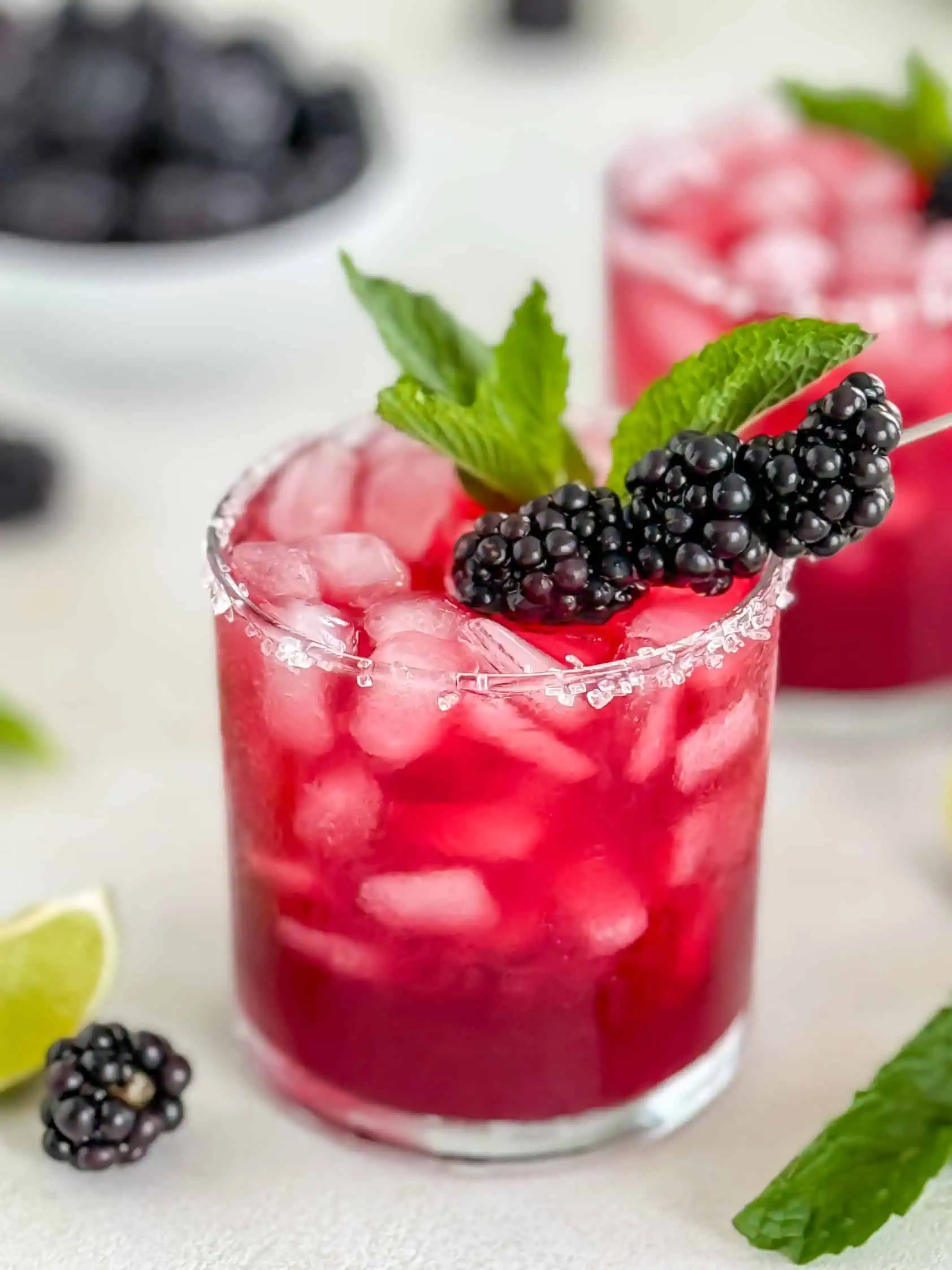 Two margaritas in glasses with sugared rims, garnished with blackberries and mint leaves, with lime wedges and blackberries around for decoration.