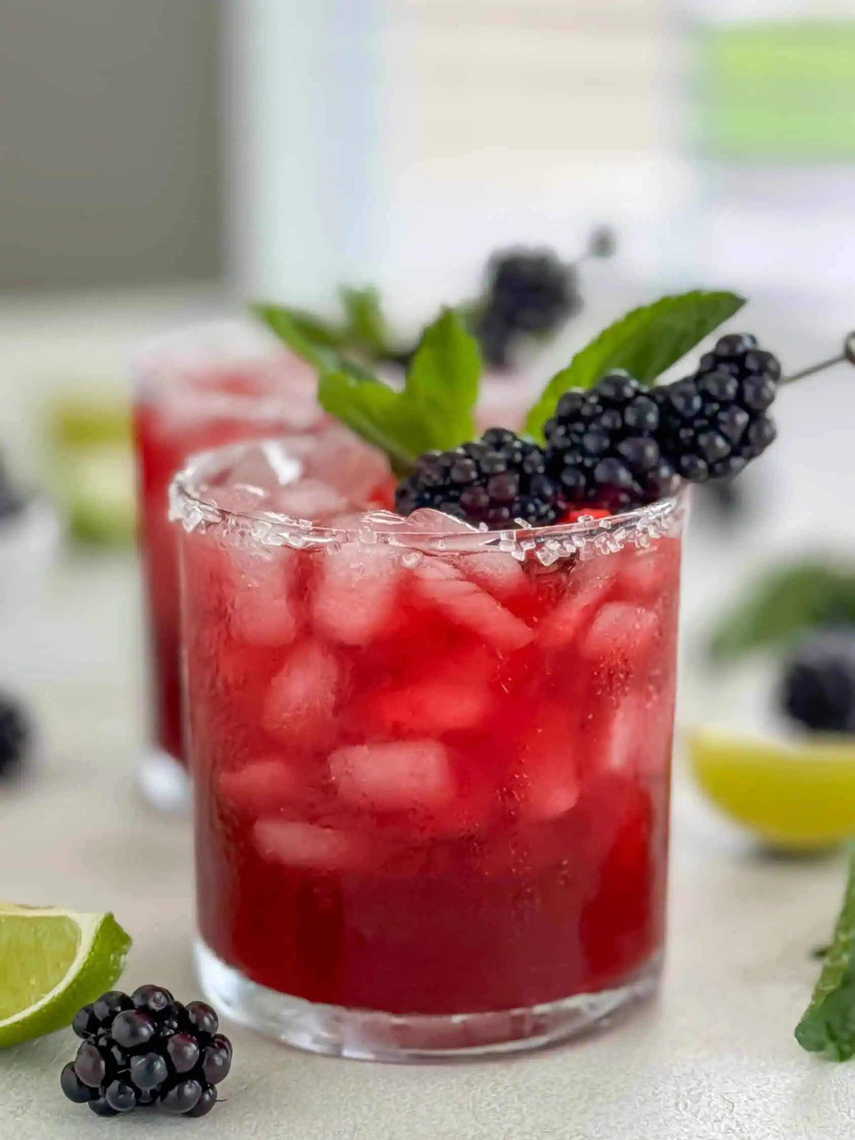 A margarita in a glass with a sugared rim, garnished with fresh blackberries and mint leaves, with additional garnishes of lime and blackberries nearby.