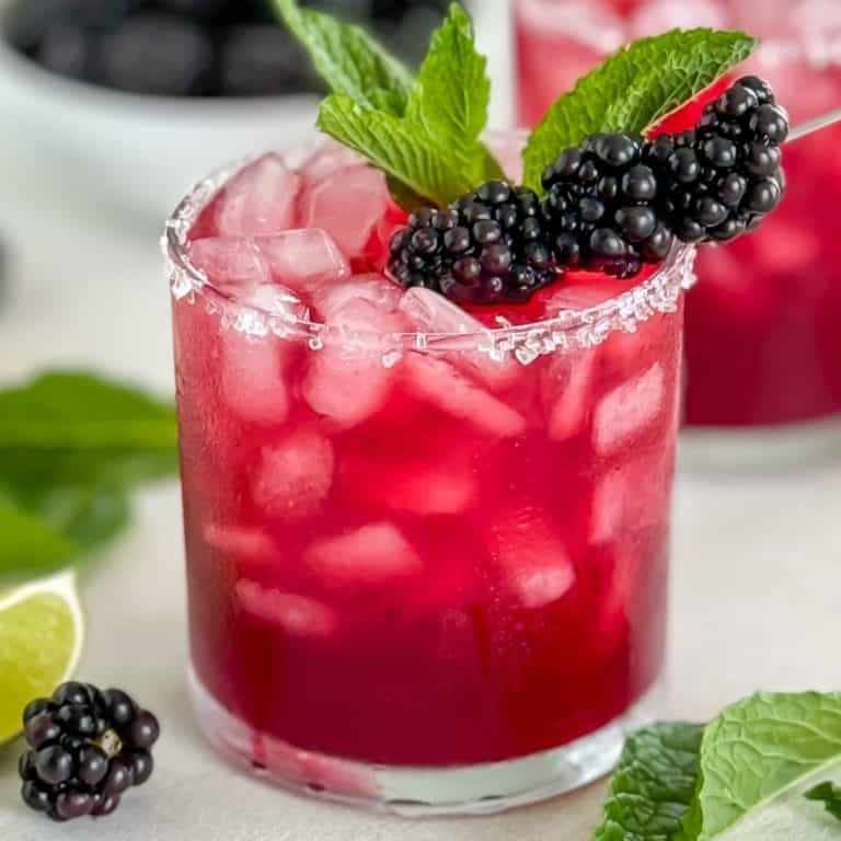 Featured image of a blackberry margarita in a glass with a sugared rim, garnished with fresh blackberries and mint leaves, with lime wedges and blackberries in the background.