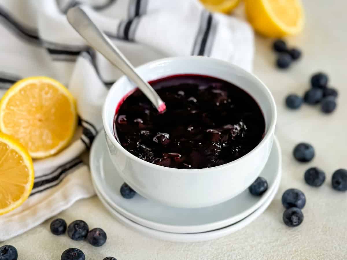 Close-up of a white bowl of blueberry compote with a spoon, fresh blueberries, and lemon halves on a light background with a striped cloth.