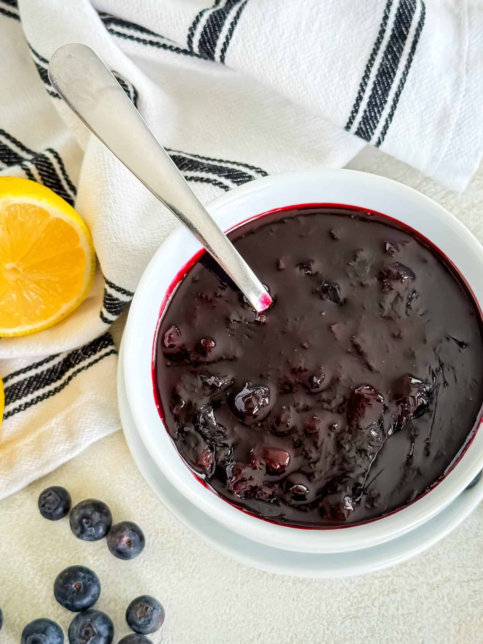 Overhead view of a white bowl with a spoon, filled with a thick, dark berry mixture, surrounded by lemon halves and fresh blueberries on a light background with a striped cloth.