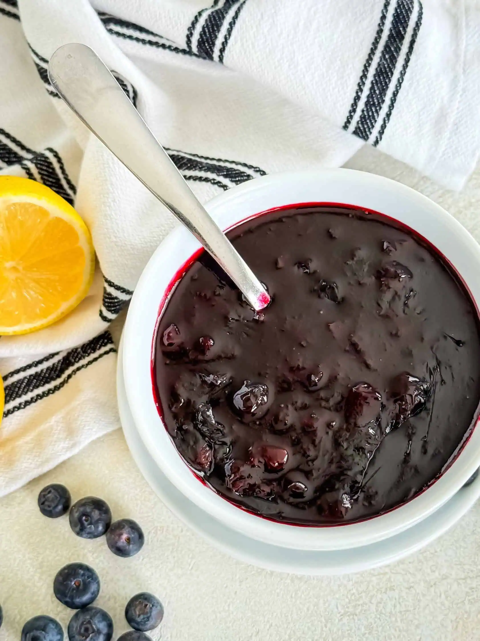 Overhead view of a white bowl with a spoon, filled with a thick, dark berry mixture, surrounded by lemon halves and fresh blueberries on a light background with a striped cloth.