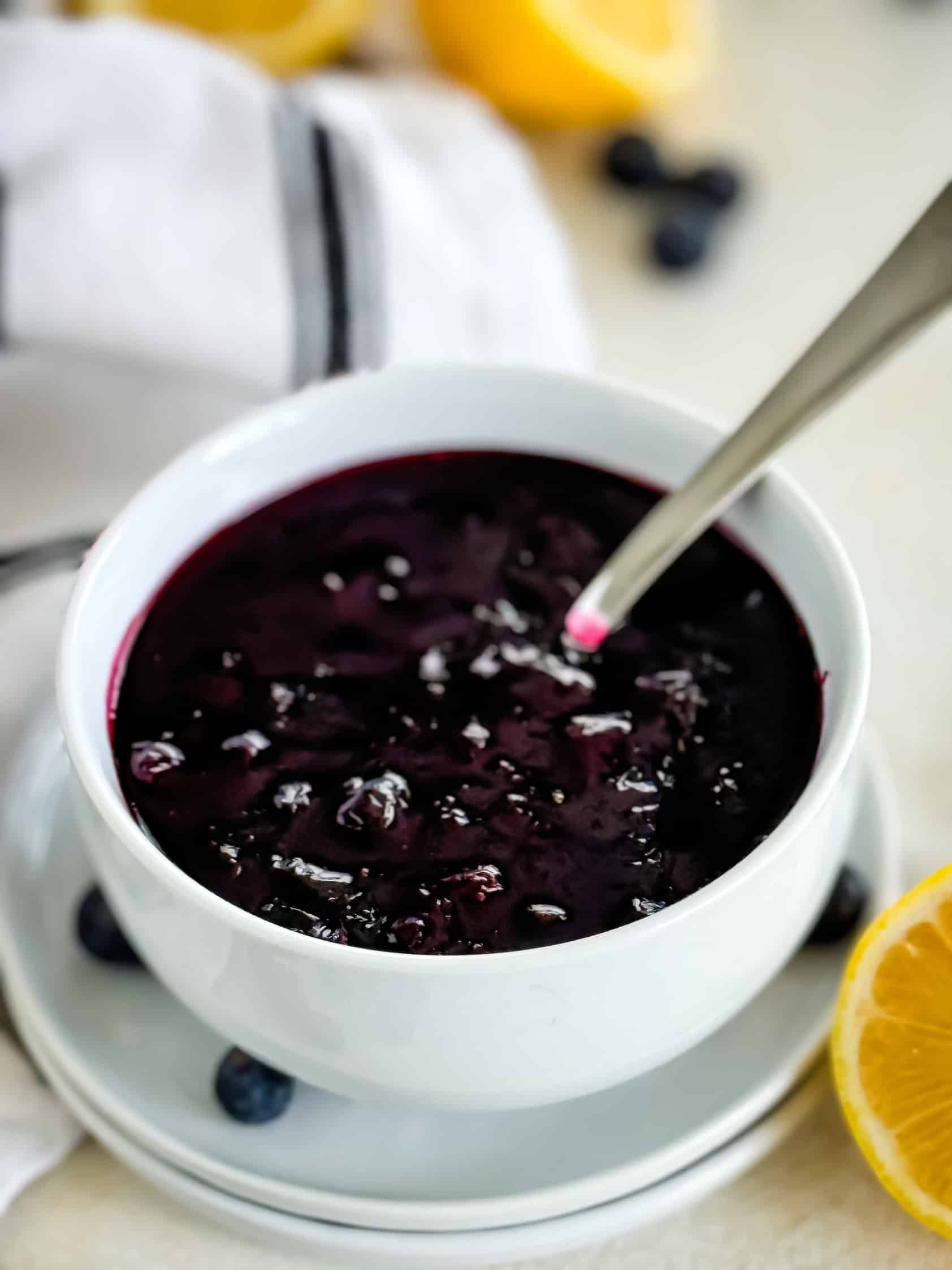 Close-up of blueberry compote in a white bowl with a spoon, surrounded by fresh blueberries and lemon halves on a light background with a striped cloth.