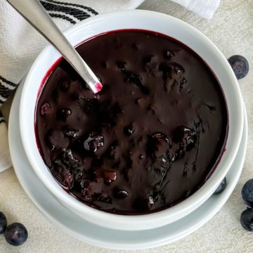 Overhead view of a white bowl with a spoon inside, filled with a rich, dark berry mixture, surrounded by lemon halves and fresh blueberries on a light background.