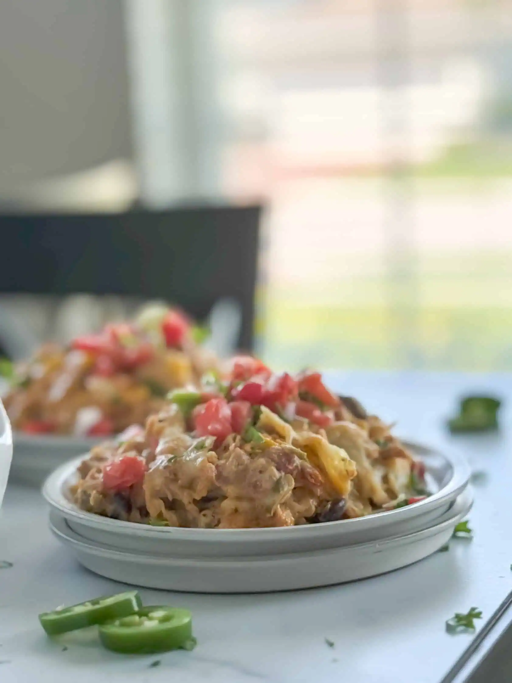 Two plates of creamy chicken taco casserole with diced tomatoes and cilantro, with the casserole dish in the background and a bright, airy dining room setting.