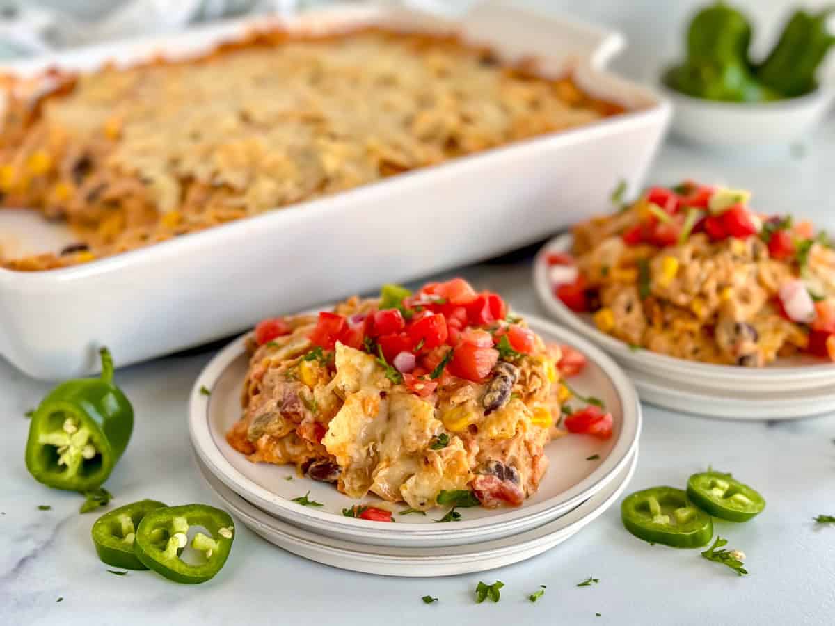 Two plates of creamy chicken taco casserole topped with diced tomatoes and cilantro, with the casserole dish in the background and sliced jalapeños on the table.