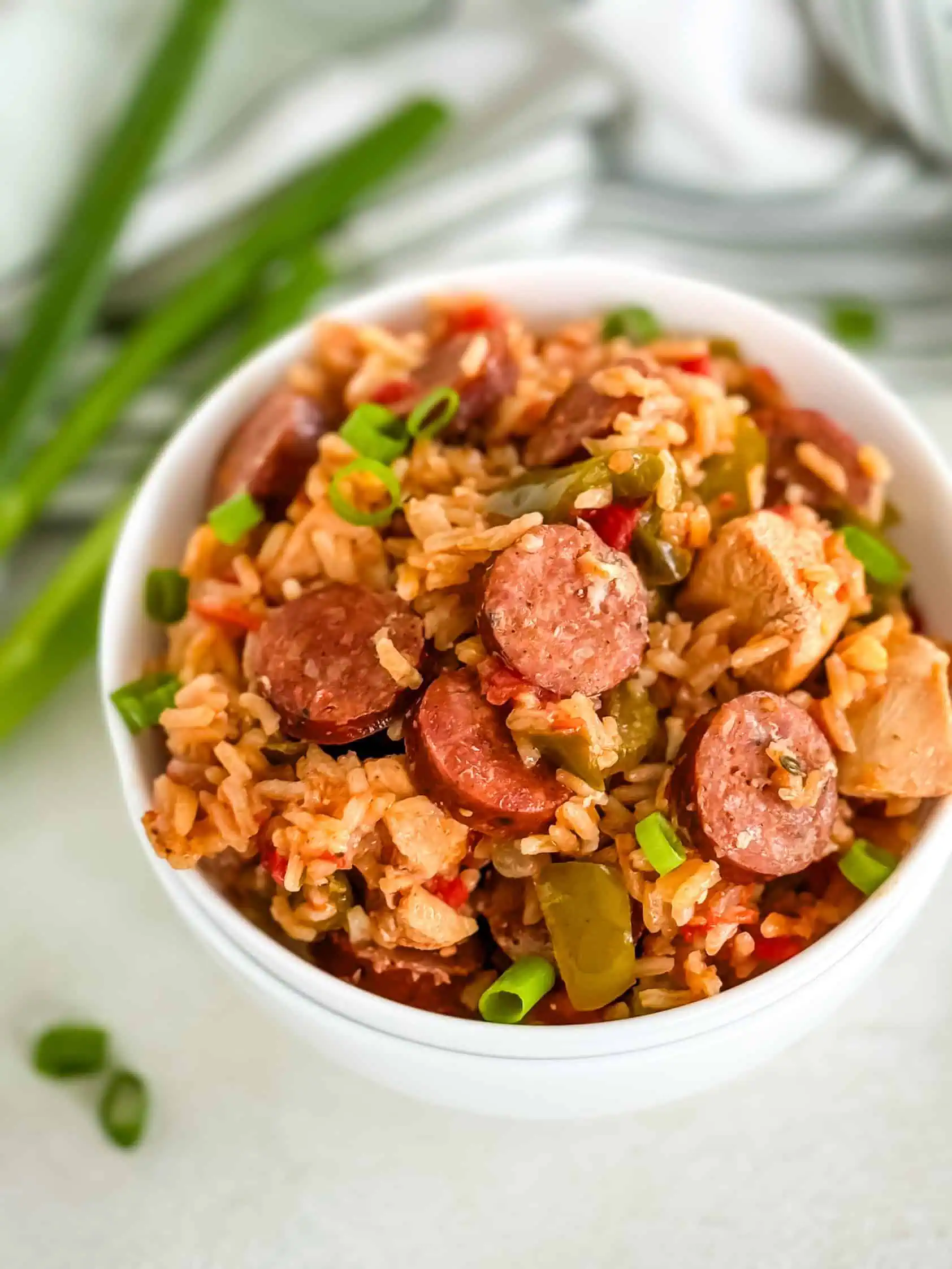 A close-up view of a bowl filled with a colorful rice dish featuring sliced sausage, diced chicken, bell peppers, and green onions.