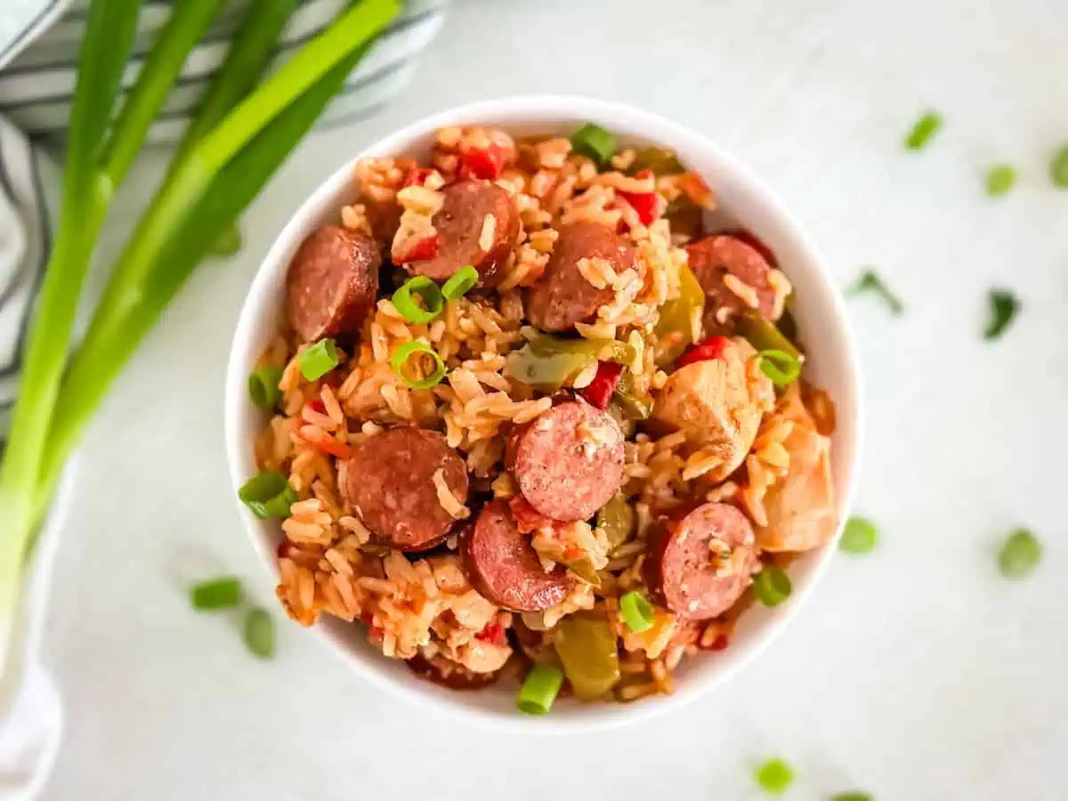 A vibrant rice dish with sausage and chicken, displayed in a white bowl, surrounded by scattered green onions.