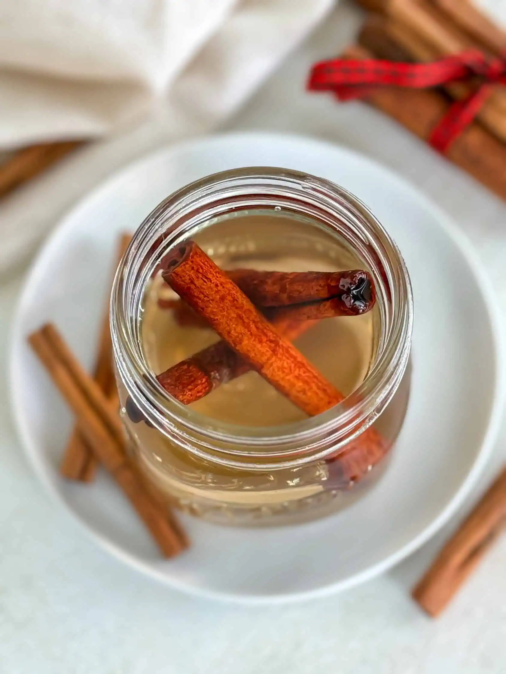 Overhead view of a mason jar filled with homemade cinnamon simple syrup and cinnamon sticks, sitting on a white plate with more cinnamon sticks in the background.