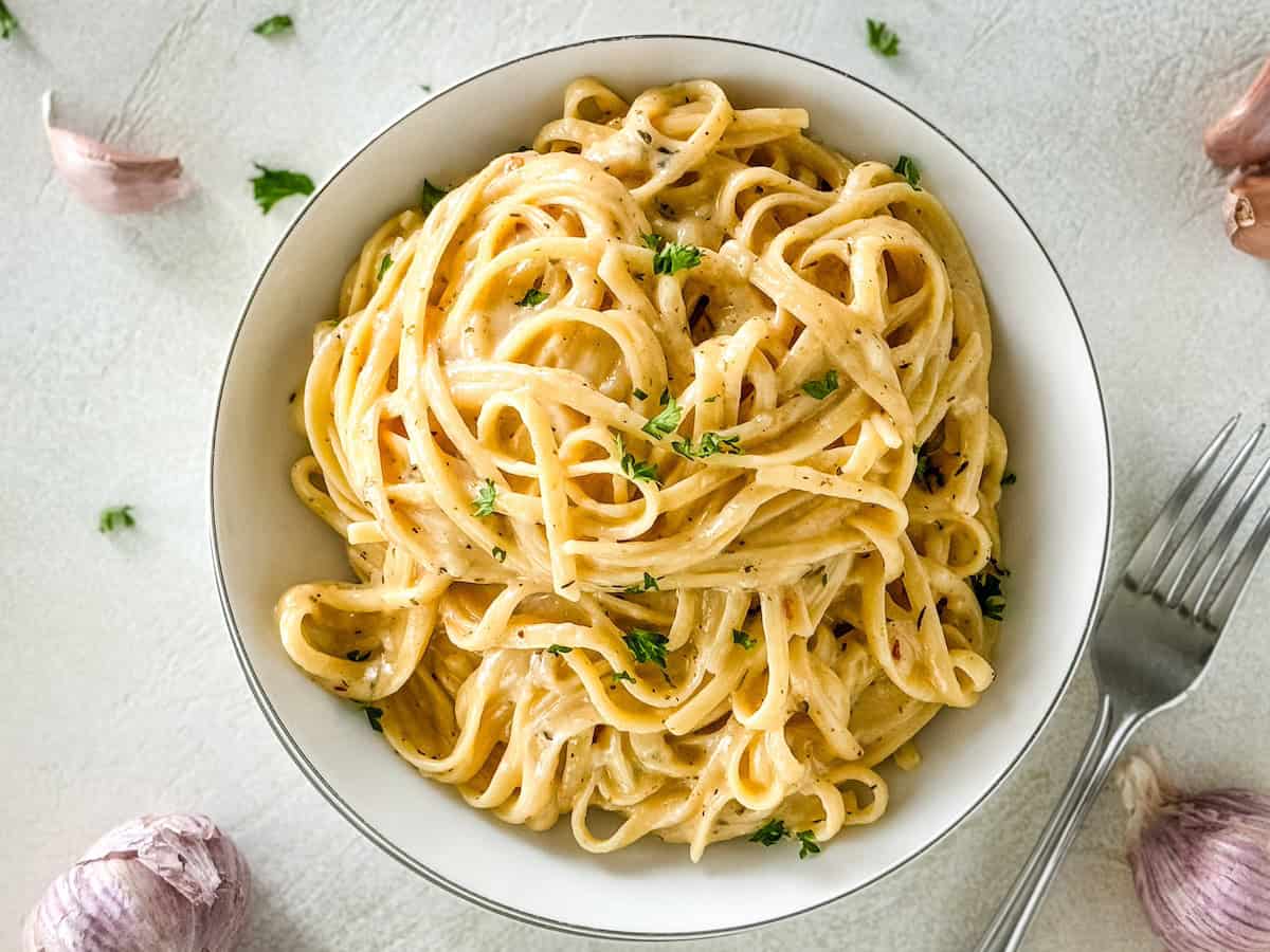 Creamy pasta in a bowl with scattered garlic cloves and parsley around.