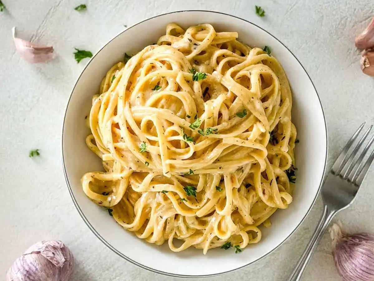 Creamy pasta in a bowl with scattered garlic cloves and parsley around.