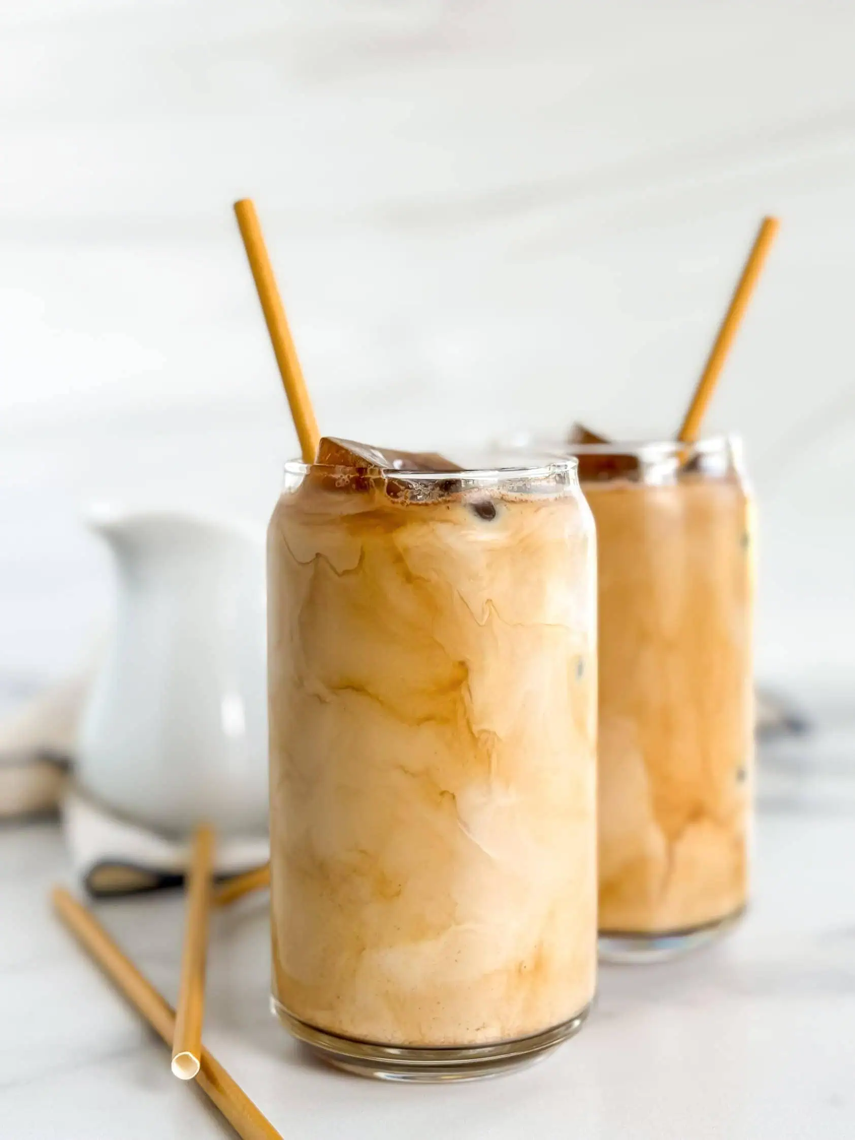 Two glasses of iced coffee with bamboo straws, with a white pitcher in the background.