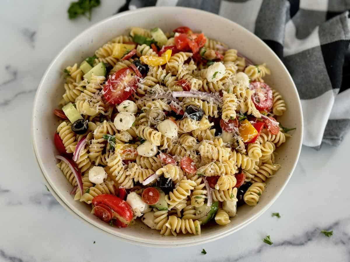 A vibrant bowl of rotini pasta mixed with colorful vegetables, mozzarella cheese balls, and black olives, served on a marble countertop.
