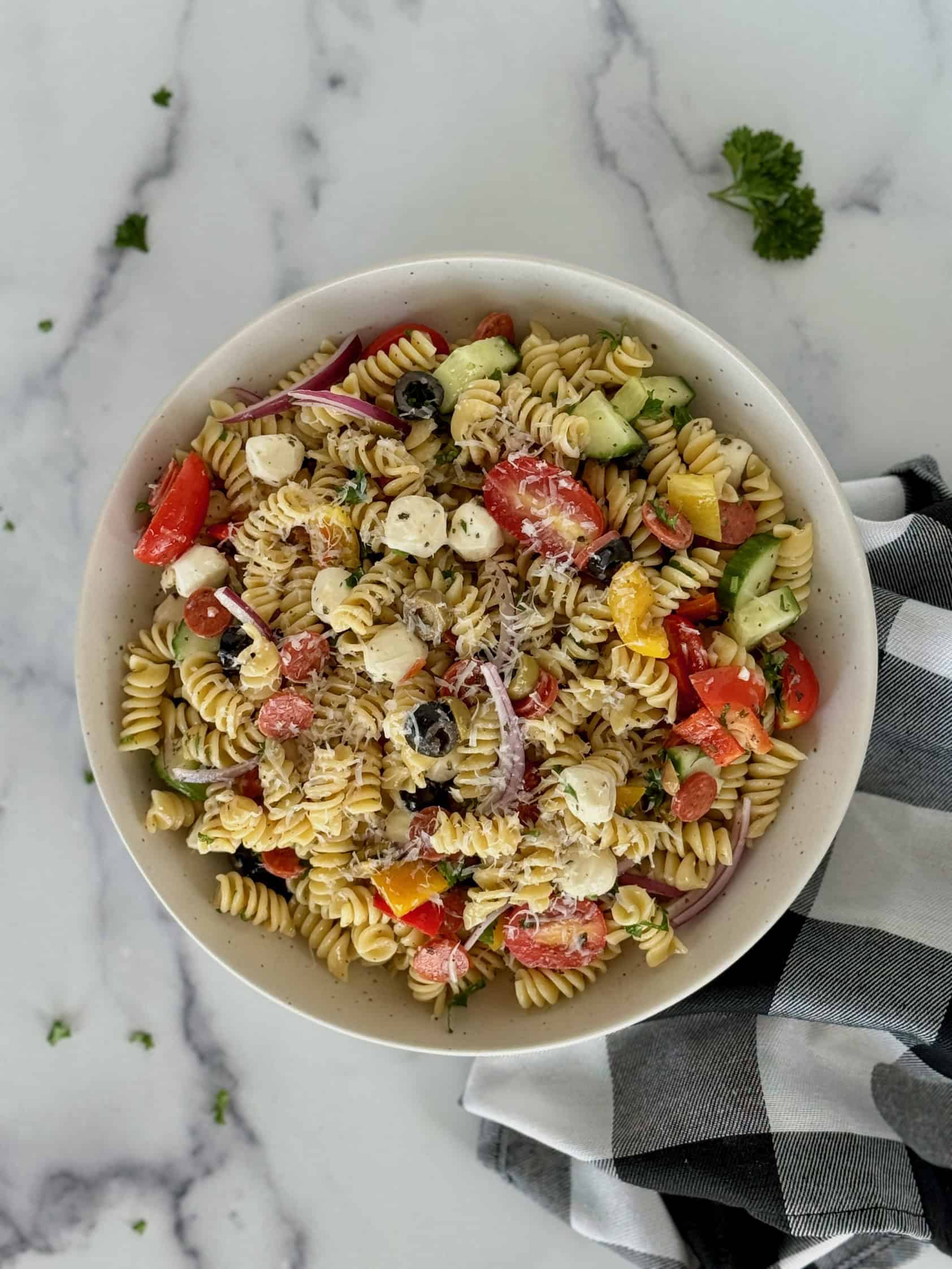 A close-up showcasing rotini pasta, cherry tomatoes, bell peppers, cucumber, red onion, black olives, and mozzarella cheese balls, garnished with fresh herbs.