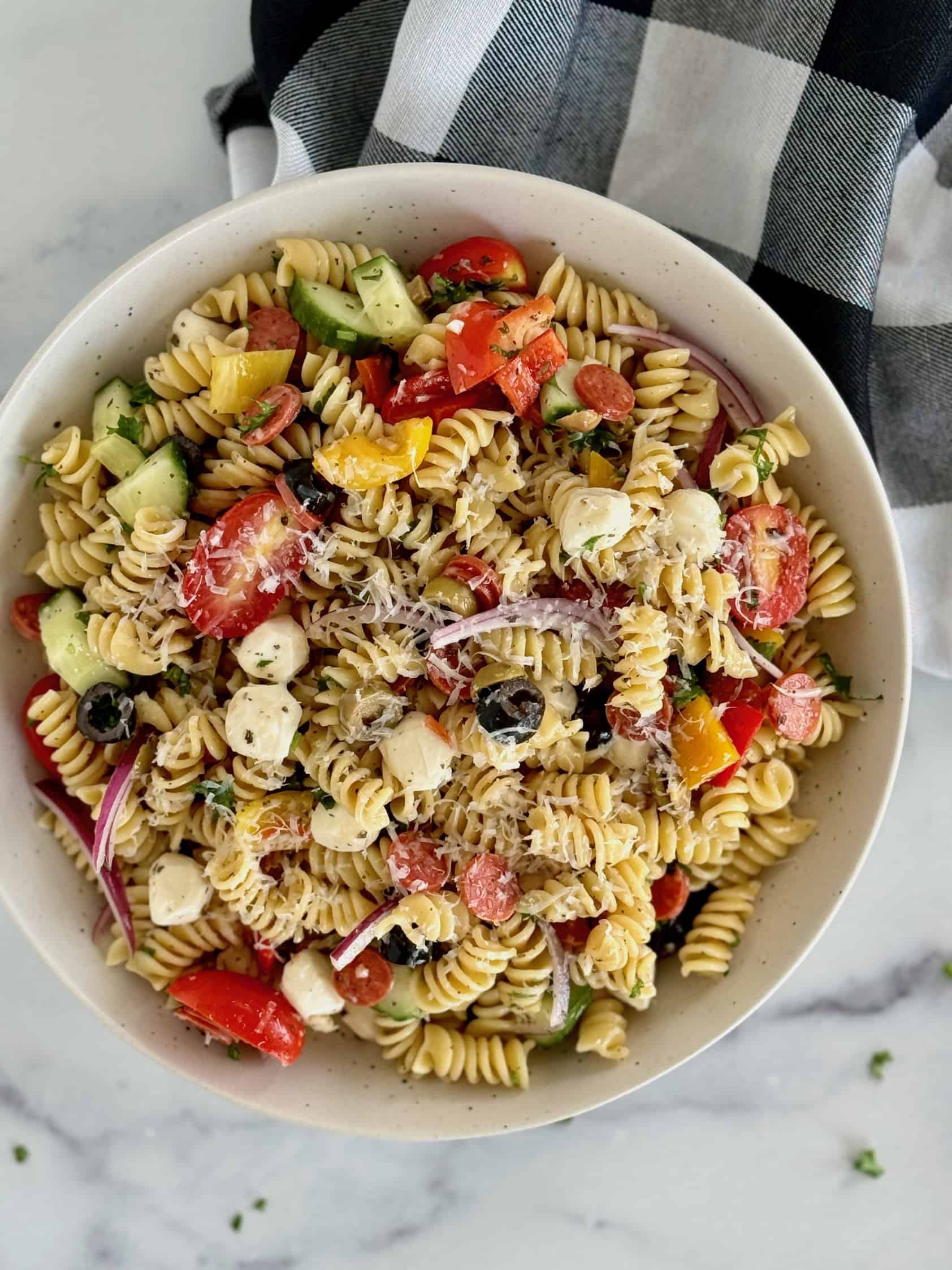 A large bowl of pasta mixed with colorful veggies, black olives, mozzarella cheese balls, and a zesty dressing, perfect for a summer picnic or bbq.