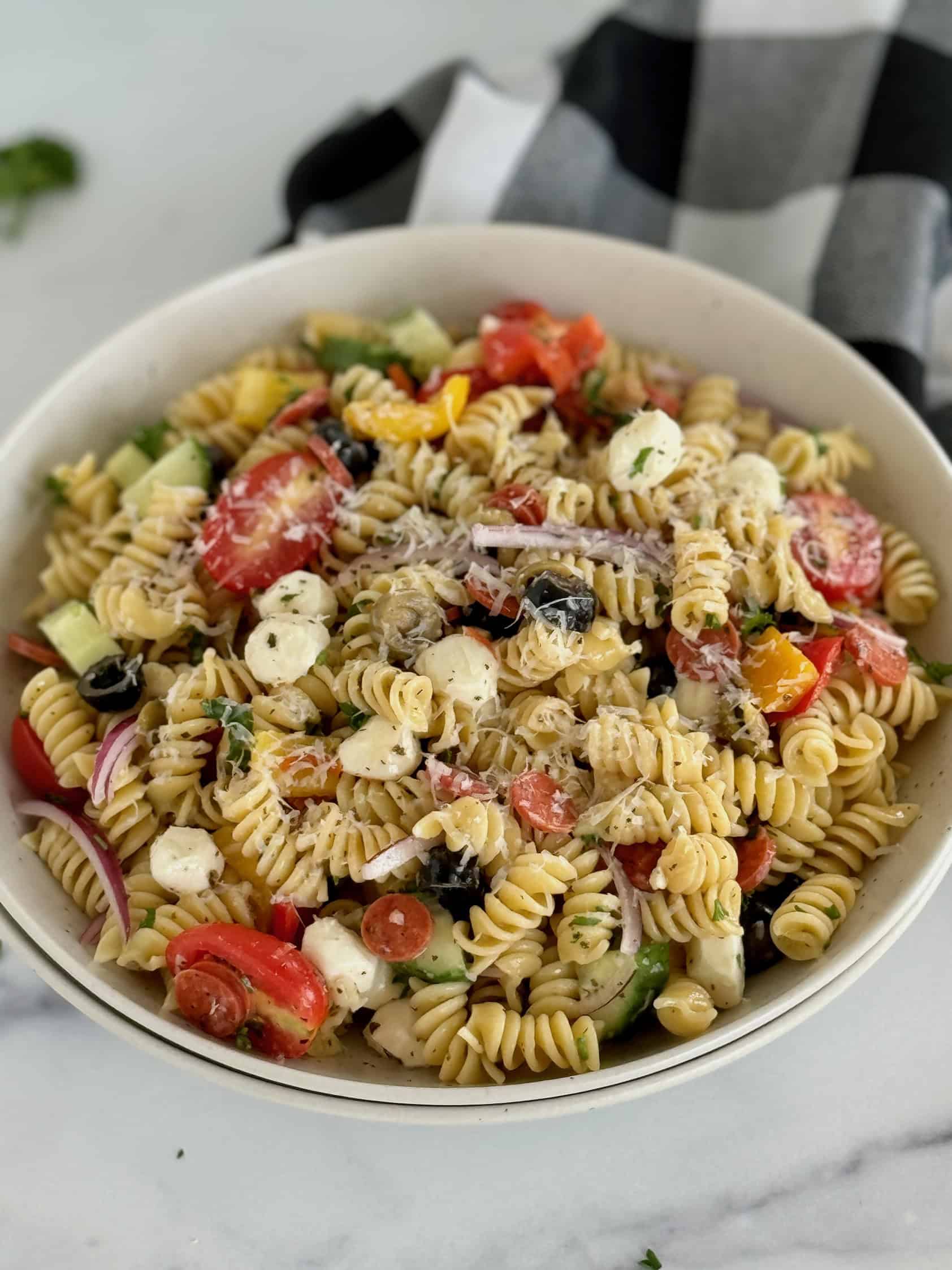 An overhead shot of a bowl featuring rotini pasta, cherry tomatoes, bell peppers, black olives, cucumber, red onion, and mozzarella cheese balls, served with a black and white checkered napkin.
