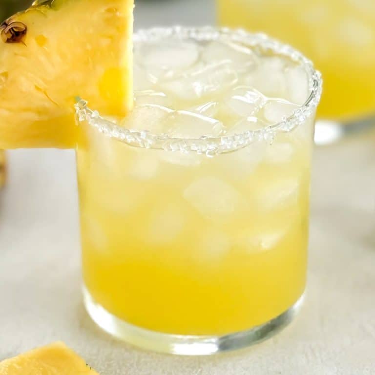 Refreshing yellow cocktail with a slice of pineapple on the rim and a textured salted rim.
