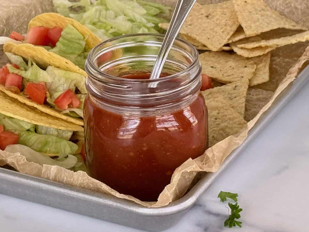 Jar of homemade taco sauce with a spoon, surrounded by tacos with lettuce and diced tomatoes, and tortilla chips on a tray.