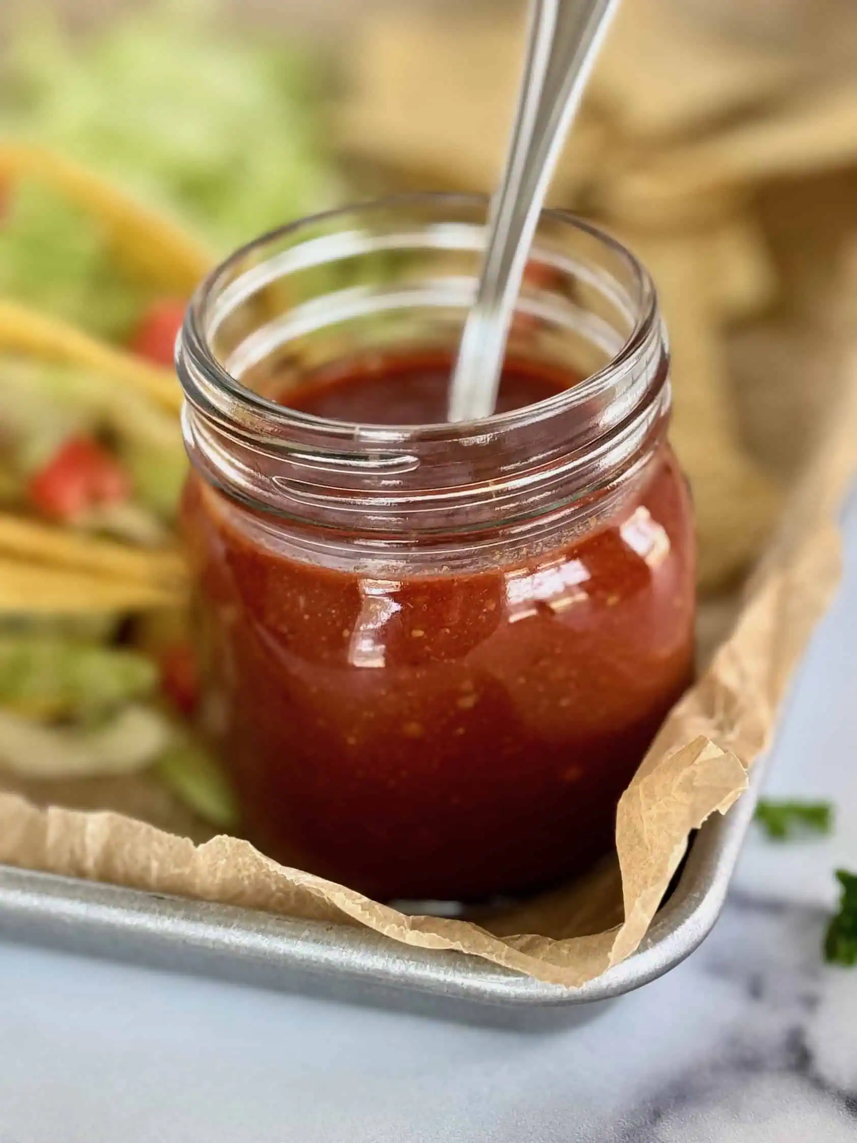 Close-up of a jar of homemade taco sauce with a spoon, surrounded by tacos and tortilla chips on a tray.