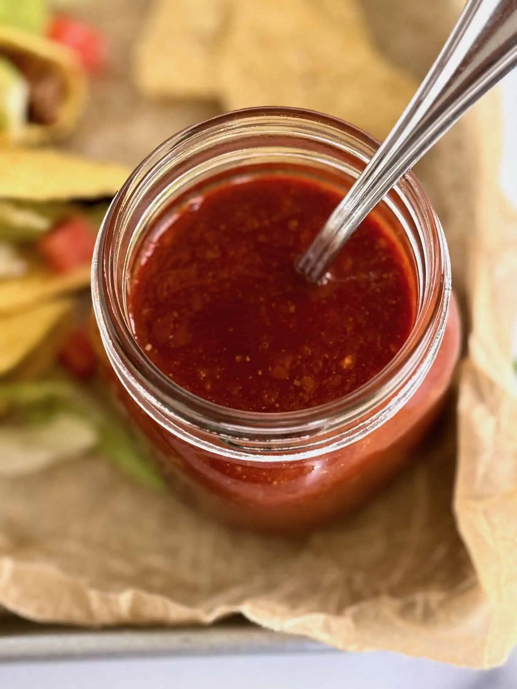 Top view of a jar of homemade taco sauce with a spoon, surrounded by tacos with lettuce and diced tomatoes, and tortilla chips on a tray.