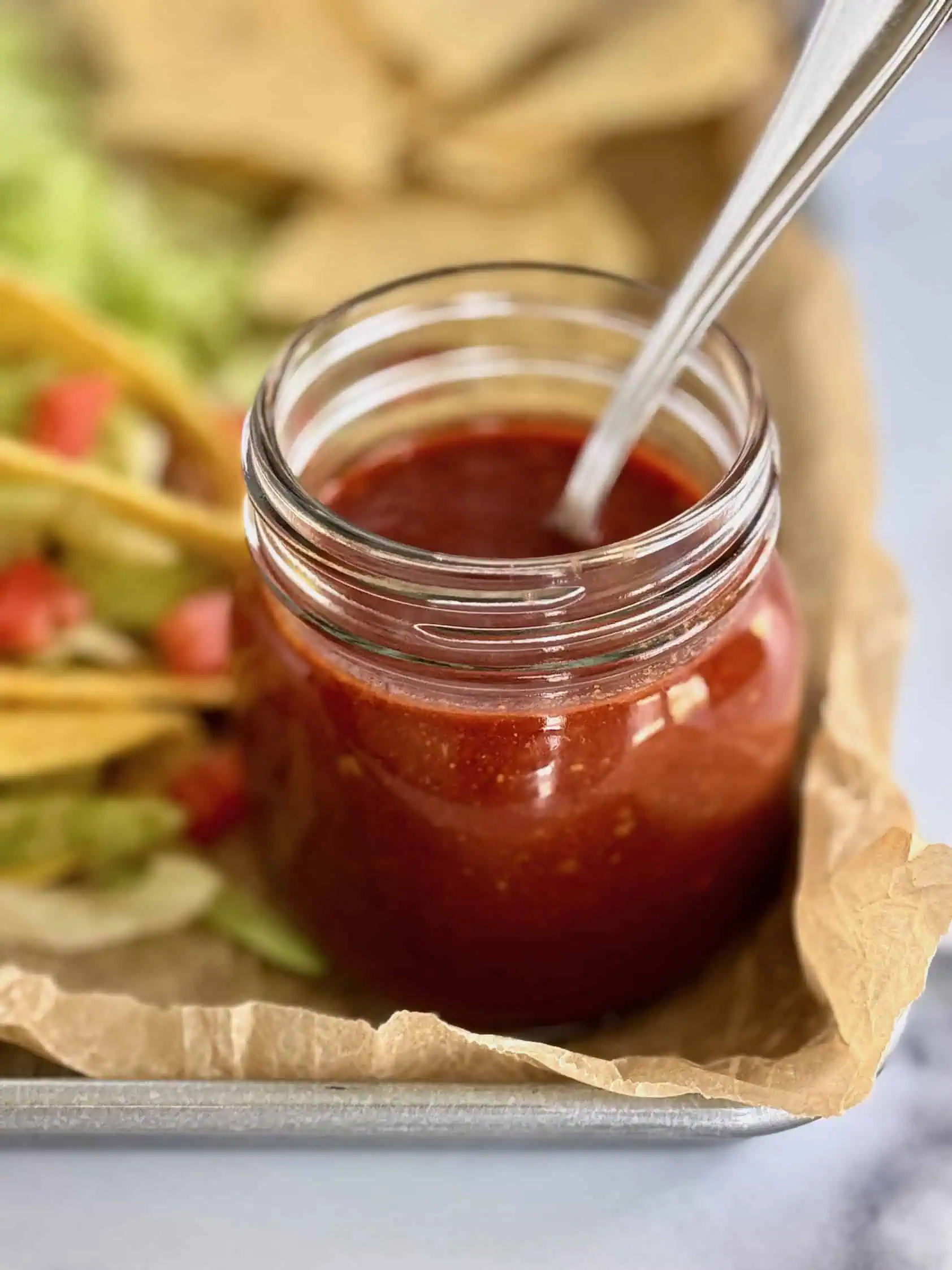 Jar of homemade taco sauce with a spoon on a tray lined with parchment paper, with tacos and tortilla chips in the background.