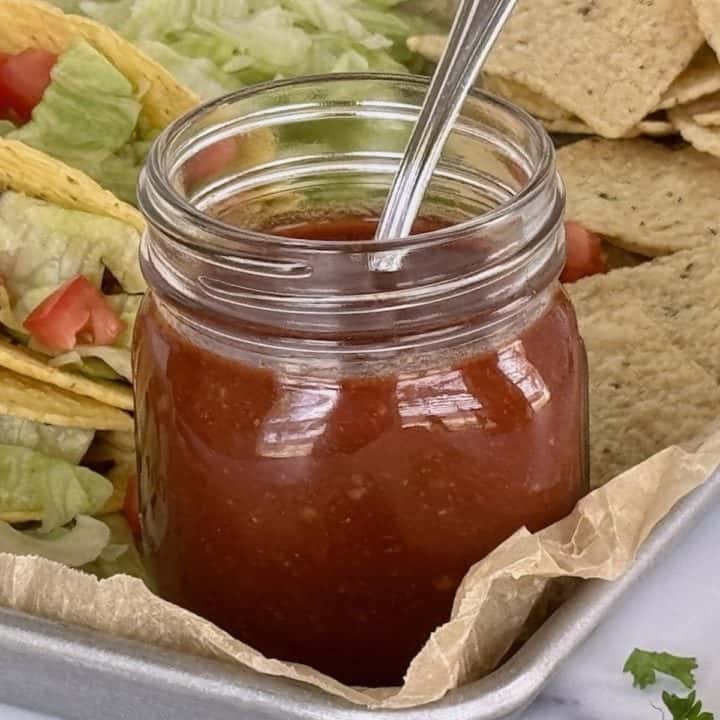 Jar of homemade taco sauce with a spoon, surrounded by tacos with lettuce and diced tomatoes, and tortilla chips on a tray.