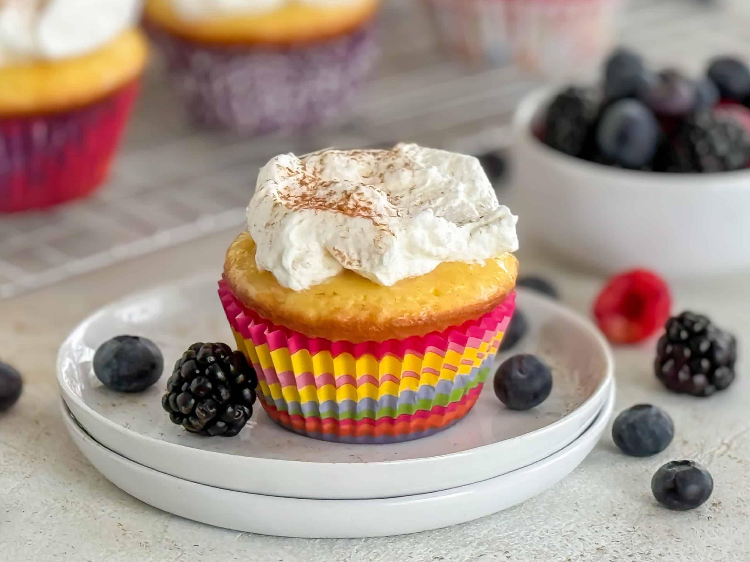 A close-up of a cupcake topped with whipped cream and a dusting of cinnamon, surrounded by fresh blackberries and blueberries on a white plate.